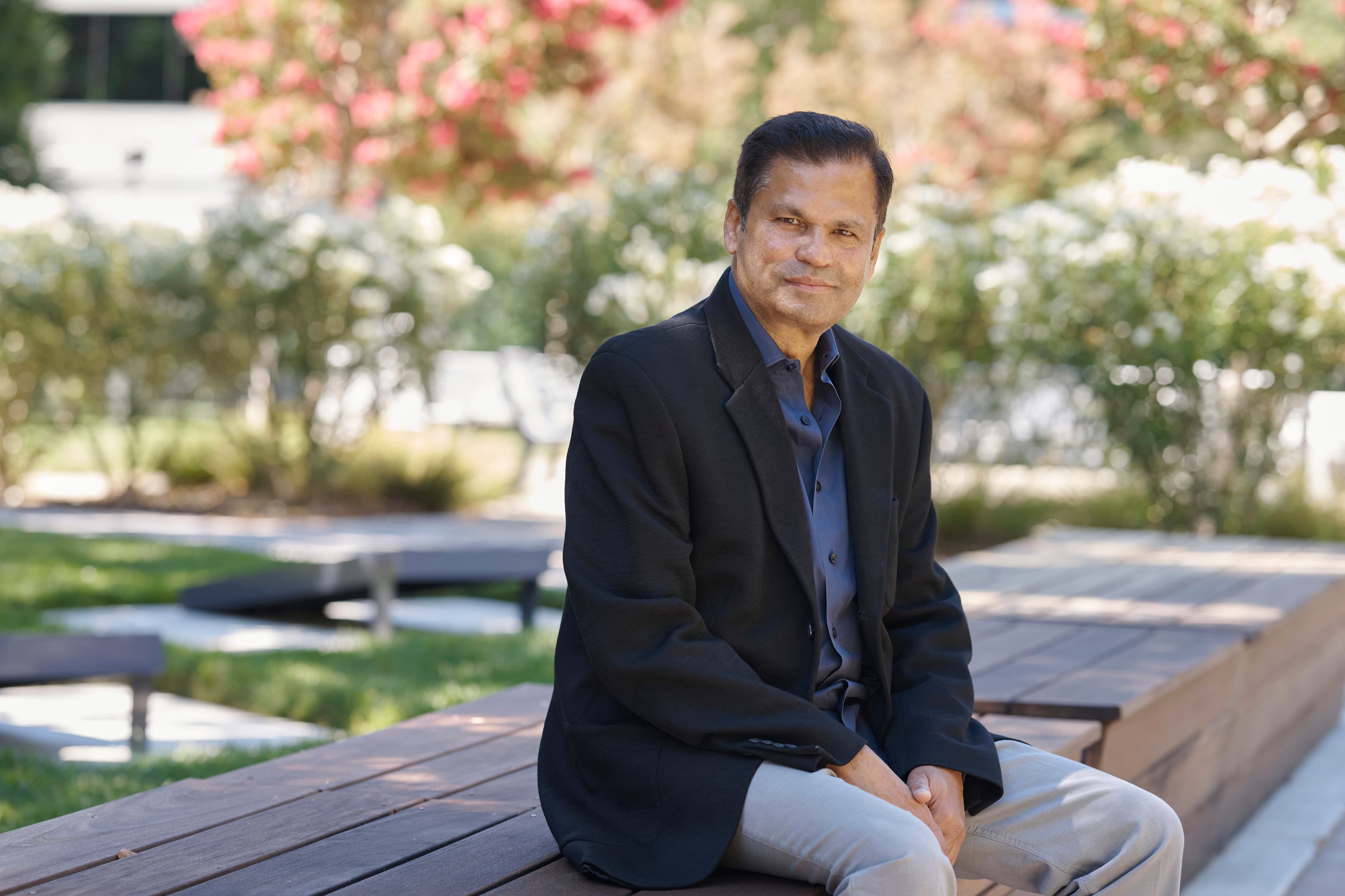 Smiling man in a black blazer and blue shirt sitting on a wooden outdoor bench.